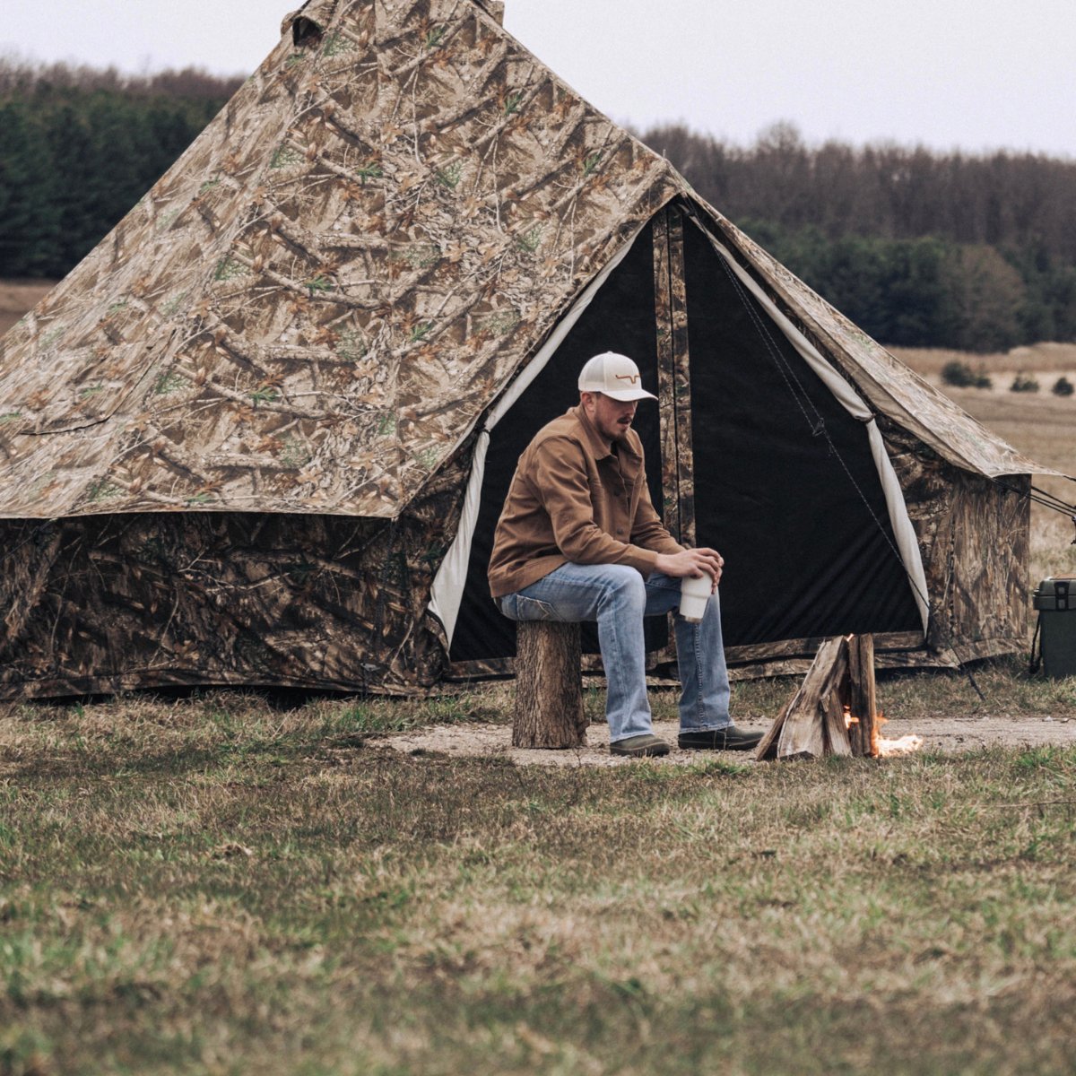 Regatta Bell Tent - Realtree Edge set up outdoors with a man sitting on a log right in front of it